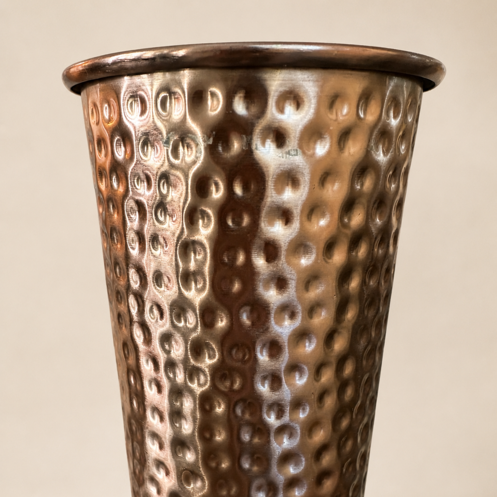 A close-up of a hammered metal decorative vase with a warm copper or bronze finish, set against a plain beige background.