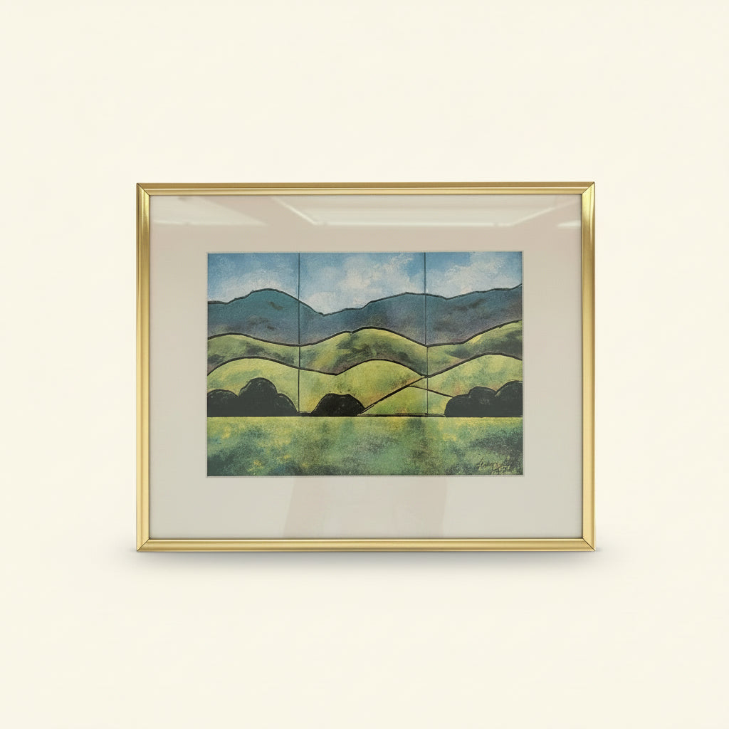 A framed landscape art print featuring green rolling hills, dark tree silhouettes, and blue mountains under a cloudy sky, enclosed in a gold frame with a white mat.