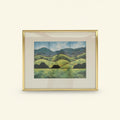 A framed landscape art print featuring green rolling hills, dark tree silhouettes, and blue mountains under a cloudy sky, enclosed in a gold frame with a white mat.
