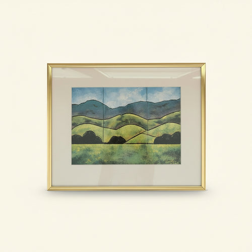 A framed landscape art print featuring green rolling hills, dark tree silhouettes, and blue mountains under a cloudy sky, enclosed in a gold frame with a white mat.