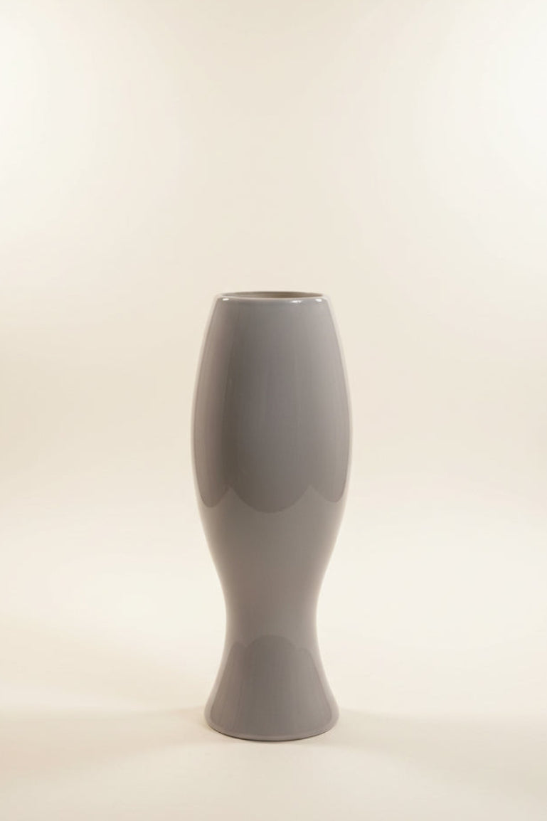 A glossy, light gray ceramic abstract sculptural vase with a flared rim and a bulbous, hourglass-like shape, displayed in a studio with ivory background, facing backward. 