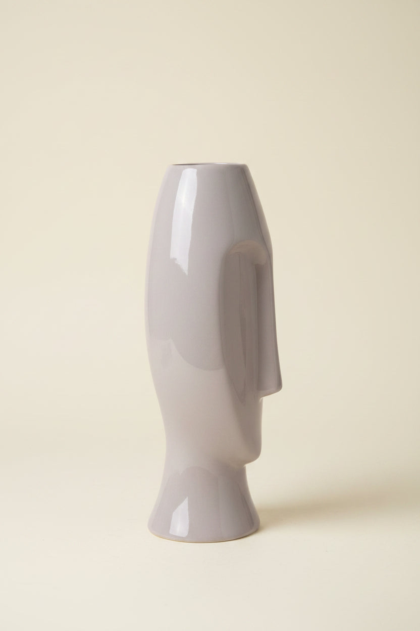 A glossy, abstract sculptural vase shaped like a stylized head, finished in a light gray color, facing left in a studio image with an ivory background.