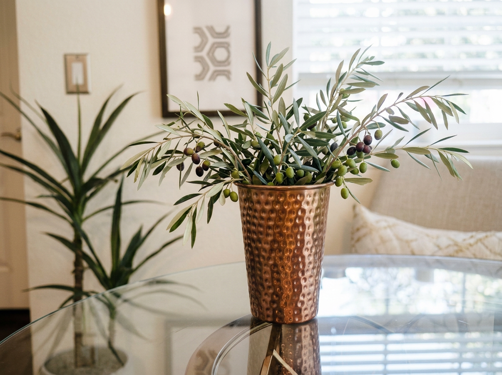 8" copper-toned vase on a glass dining table in a bright dining room styled with olive branches. 