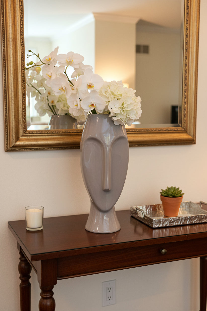 A glossy, modern sculptural ceramic vase shaped like a stylized face, in a light gray color, displayed on a glass-topped table in a living room setting.
