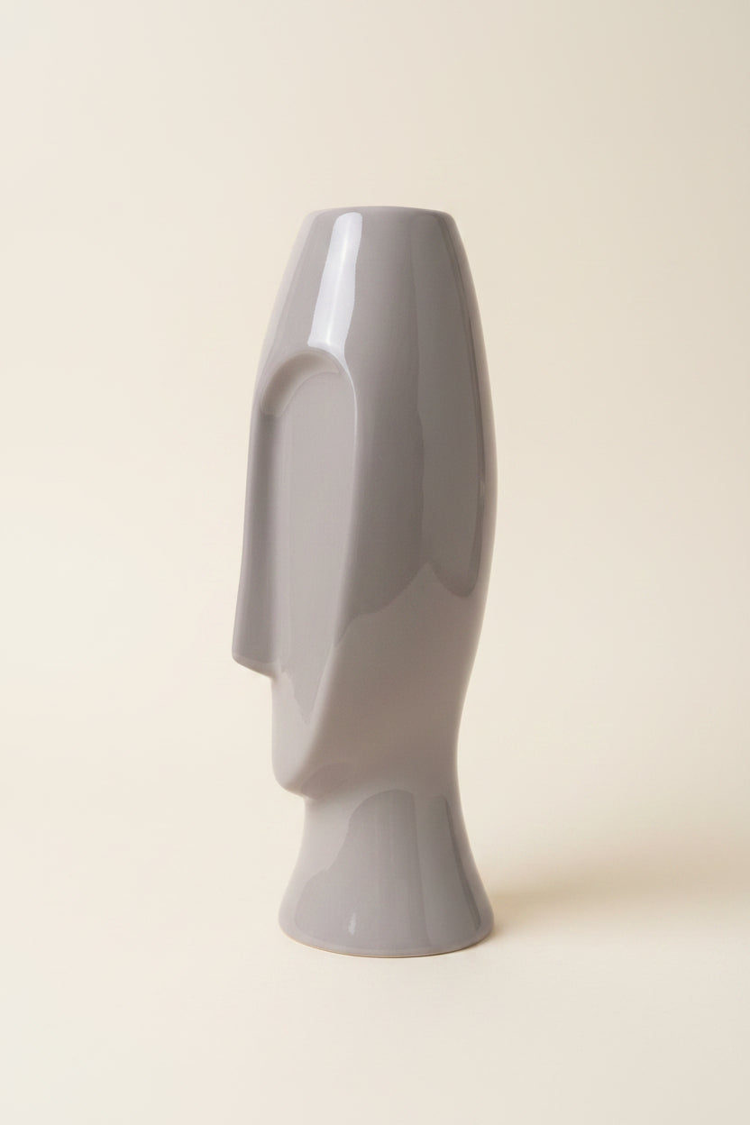 A glossy, abstract sculptural face vase in a light gray color, displayed in a studio background facing right. 