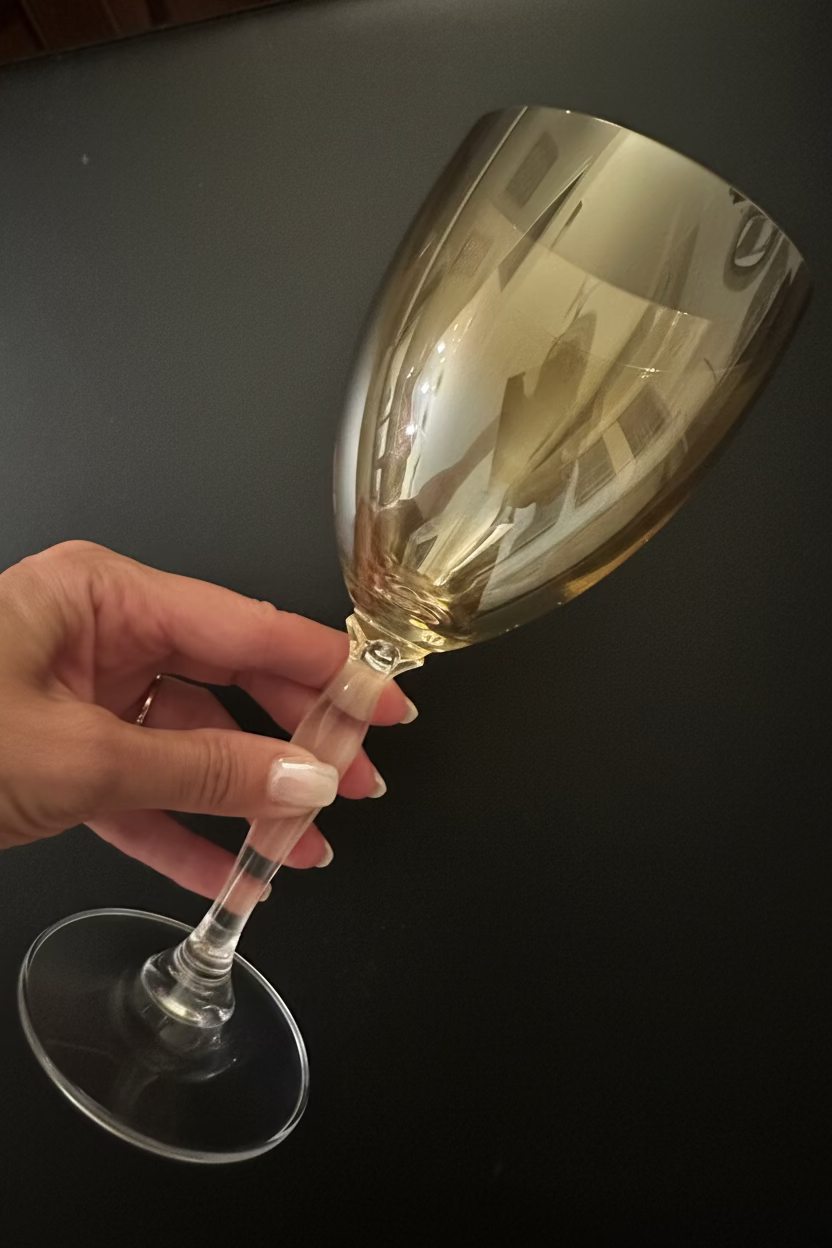 A hand holding a clear wine glass with a reflective gold-toned tint against a dark background.
